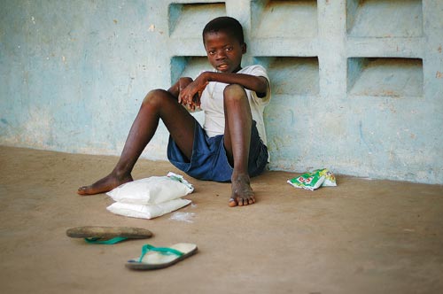 A young boy sits on a brown cement floor, leaning against a pale blue cement wall, legs sprawling out before him. He's taken off his sandals. Around him are several bags full of white powder. He looks directly at the camera.
