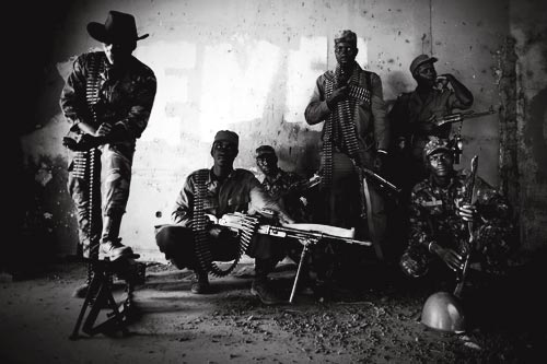 Half a dozen soldiers pose for a photograph. They are heavily armed, half of them with ammunition slung over their shoulders. One wears a cowboy hat, shielding his eyes from the camera. All are wearing military uniforms, some camouflaged.