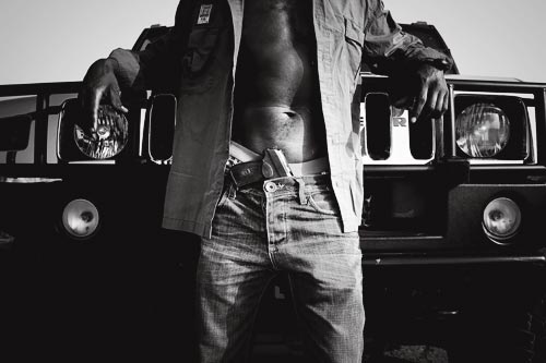 A man stands in front of a Hummer, posing as if for the cover of a rap album. He is photographed from the chest down to the knees. His button-down shirt is open in the front, revealing high-riding boxers and a handgun stuffed into the waist of his stylish jeans. The grill of his vehicle is polished to a high shine.