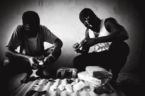A pair of masked men process bricks of drugs, breaing off pieces and wrapping them up for sale on a smaller scale. They each wear ski masks. One of them wears a t-shirt promoting Ohio Girls Basketball.