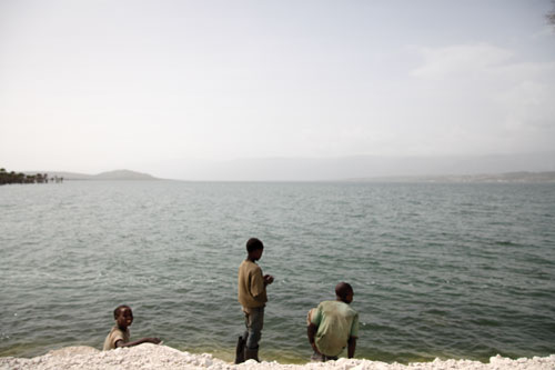 Three young Hatian boys stand on a rocky white beach above the ocean.
