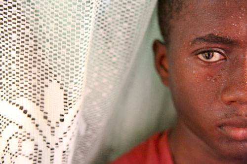 A closeup of a young Hatian boy in a red t-shirt. He is flanked on one side by a lacy white curtain.