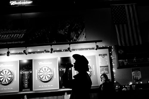 Nick, silohuetted against an illuminated wall of dart boards in a bar.  He is wearing a cowboy hat and singing into a microphone.  A woman looks on in the background.