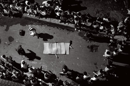 Seen from above, a circle of people crowd around a body which lies on the pavement covered by a blanket.