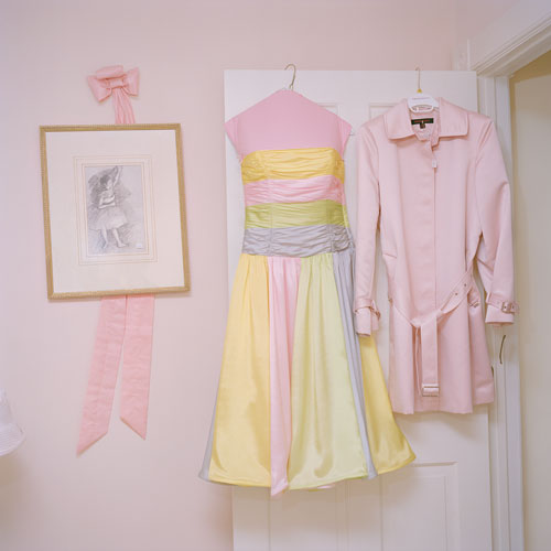 A close cropped shot of the open door of a pink room.  There is a pink ribbon on the wall behind a framed picture, and from the door hangs a pink and yellow sun dress on a hook and a pink blouse with matching jacket.