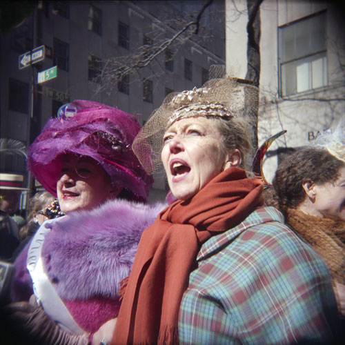 A close-up shot of two women in in hats, scarves and sashes standing on a bright, sunny New Your street.