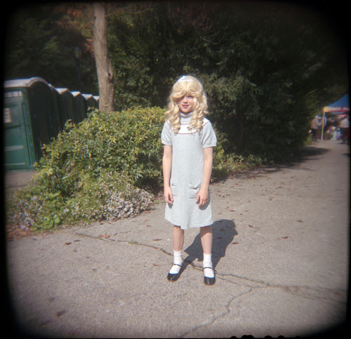 A young blonde girl in a blue sun dress standing, facing the camera, in the middle of a sidewalk.