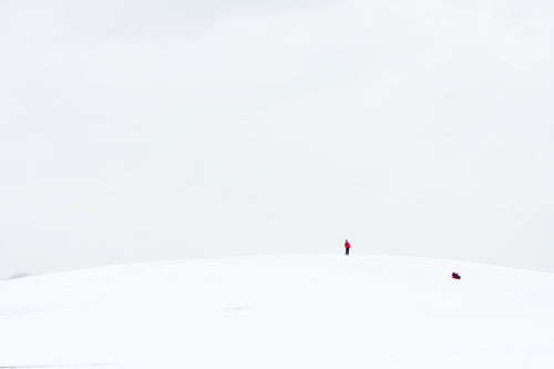 A nearly full-white image of a hill covered in snow.  On the hill, far enough away to be tiny, is a man in a red jacket.