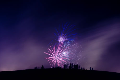 The hill, a black mound of a siluhuette against the night sky, littered with the siluhuettes of people seated, watching a colorful fireworks display in the distance.