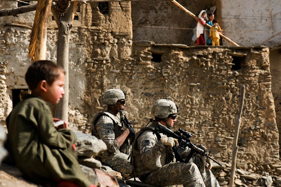 A pair of U.S. soldiers sit down on a steep hillside in a village. An Afghan boy sits nearby, and a woman and her children watch from afar.