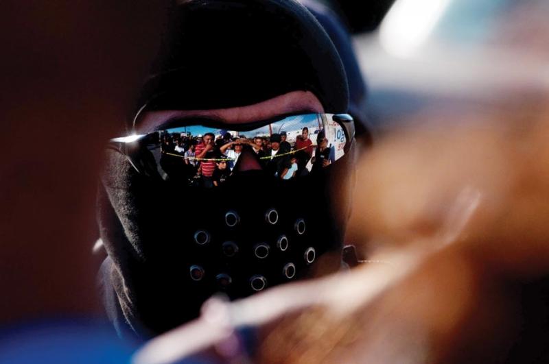 A surging, angry crowd reflects in the sunglasses of a masked police officer.
